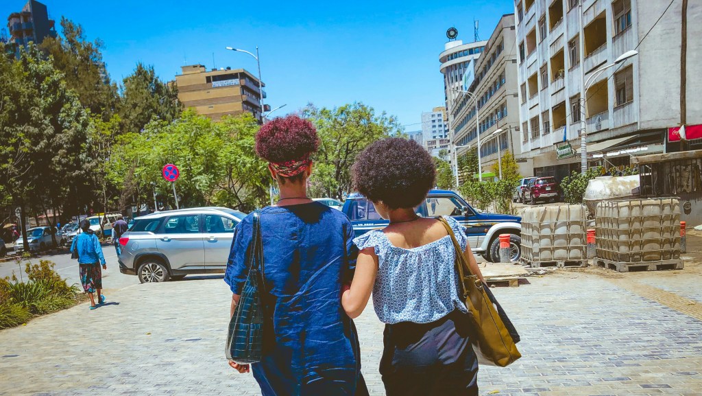 Two women stroll together along a city street, with shops and pedestrians visible in the background.